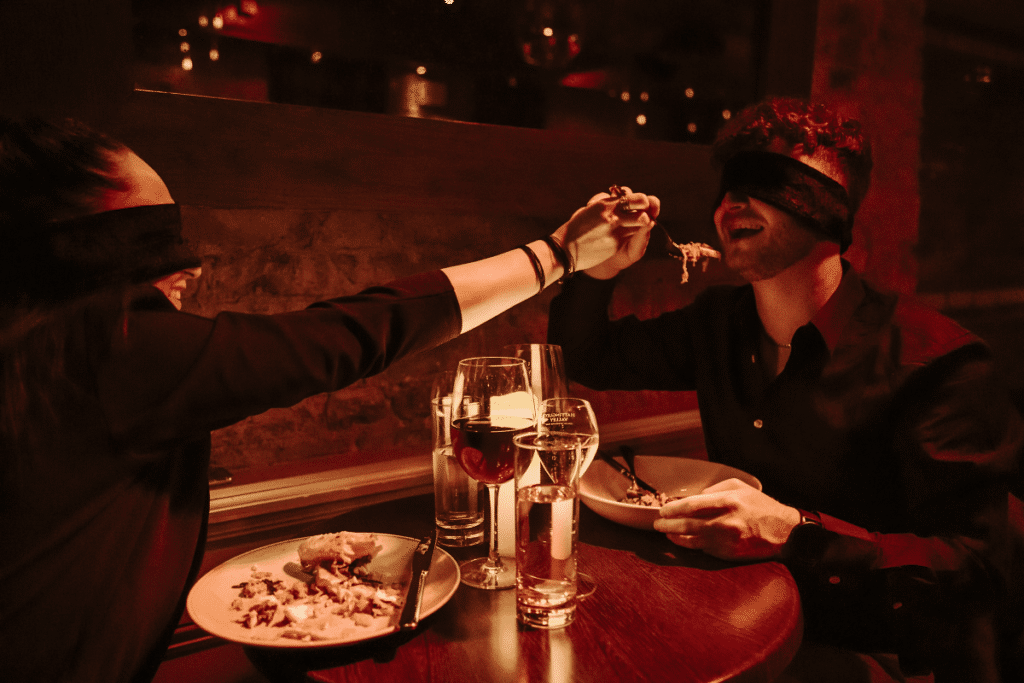 A couple holding hands and laughing while wearing blindfolds at a Dining in the Dark experience.