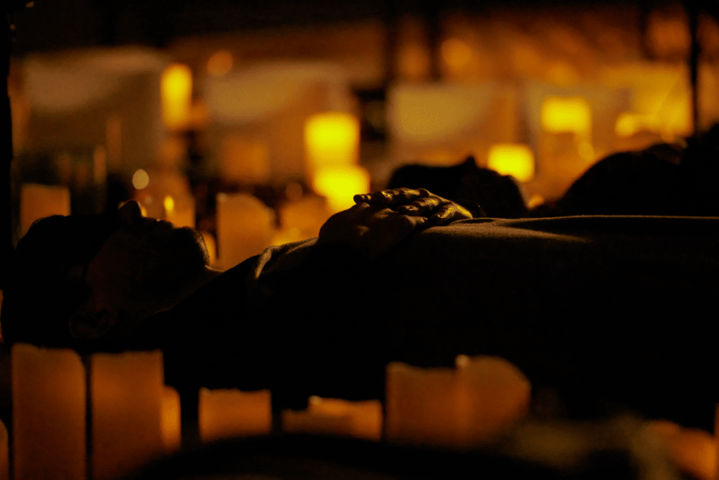 The silhouettes of two people laying on their backs and resting their hands on their chests while surrounded by countless candles