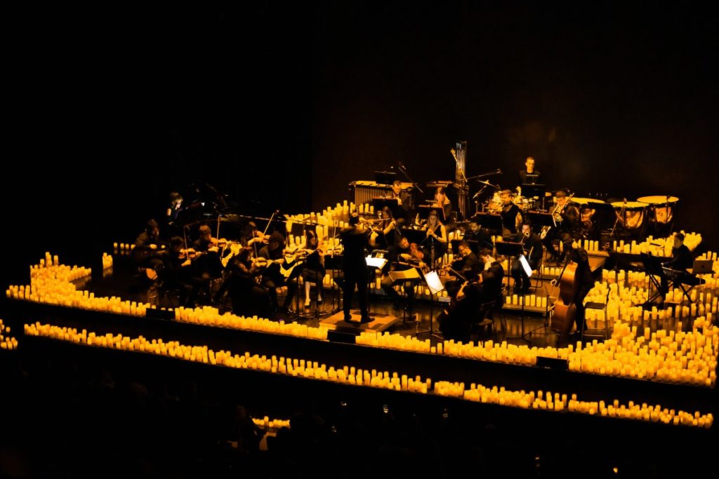 A wide shot of an orchestra performing on a raised stage fully surrounded by candles