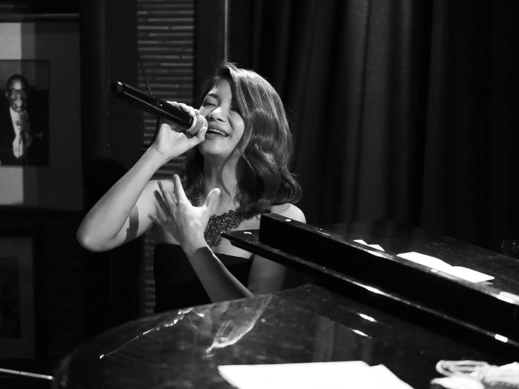 Jazz singer Natasha Patamapongs sings soulfully into a microphone whilst seated at a piano.